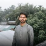 A young South Asian man stands outdoors with lush greenery in the background, exuding calmness.